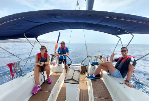 Three people on a sailing yacht, one is helming (steering) and the other two are sitting relaxing under a blue canopy