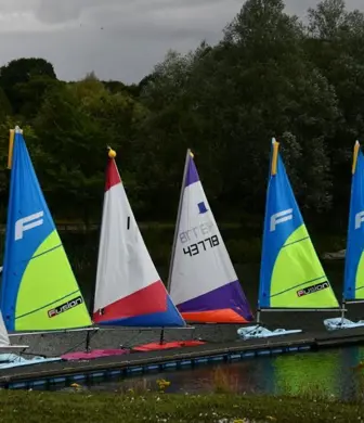 A moored line of different dinghies with different coloured sails along a jetty