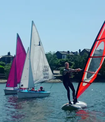 Two people sailing small sail boats alongside a man windsurfing on the water on a bright day