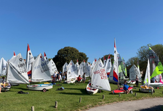 A large group of sailing dinghies on trollies gathered on a green for a Junior Regatta