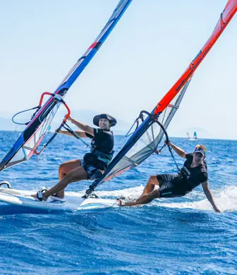 Two men enjoying skimming across beautiful blue seas on windsurfs while one of them is leaning out and dragging his hand through the water at Watersports Nomad, Vassiliki, Greece