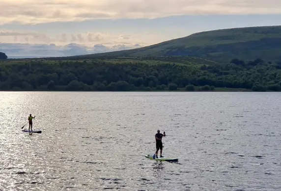 Two people on stand up paddle boards against a stunning hilly countryside background at the Watergrove Windsurfing Club, Watergrove Reservoir, Rochdale