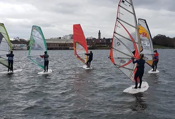 A group of five people standing on windsurf boards holding the sail up learning to windsurf at Midland Sailing Club, Birmingham