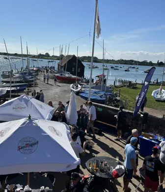A Regatta taking place with pub style umbrellas up, groups of people chatting and boats on the water in the background