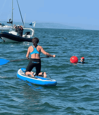 A paddle boarder kneeling on their board, alongside a swimmer in the water with a rib in the background on a sunny day