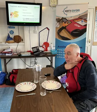 A man sat at a table laid for dinner pictured holding his throat with his mouth open and his eyes popping out during a first aid demonstration/practical at North Oxford Marine Training, Grimsby Reservoir, Banbury
