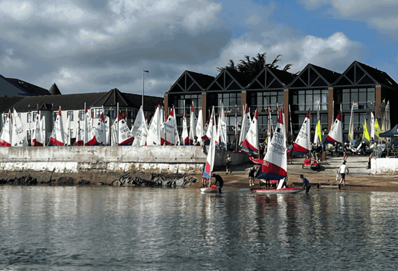 A very large group of dinghies with their sails up on the shore before their club house building