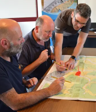 A desk with two students sat listening while an instructor demonstrates navigational techniques using charts and equipment at North Oxford Marine Training, Grimsby Reservoir, Banbury