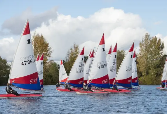 A group of small sailing dinghies with red and white sails racing on the water with trees in the background