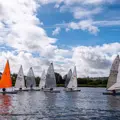 A group of Laser dinghies on the water, one with an orange sail, the rest are white with lots of white clouds in the sky