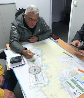 Three men around a desk plotting their journey using charts and navigational equipment