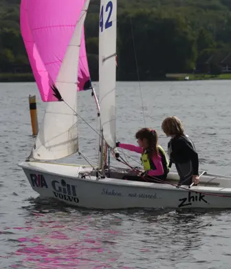 Two children sailing a Laser dinghy with a mainsail, jib and bright pink spinnaker. There's a kayaker in the distance on the water too