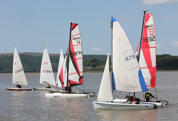 A group of dinghies sailing on the water on a sunny date with the shore in the background