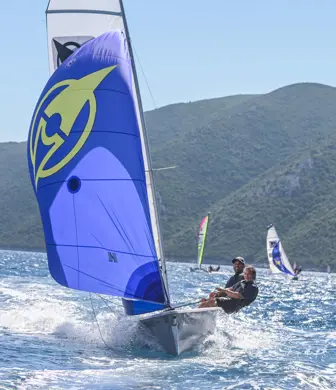 Two men sailing a small sail boat with a white mainsail and blue spinnaker on a very sunny day at Watersports Nomad, Vassiliki, Greece