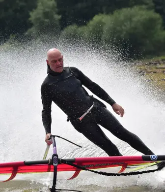 A man leaning towards the water and lowering the sail of his wind surf with water spraying out behind him at Watergrove Windsurfing Club, Watergrove Reservoir, Rochdale