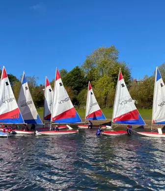 A group of eight dinghies sailing with red, white and blue sails near to the shoreline at Midland Sailing Club, Birmingham