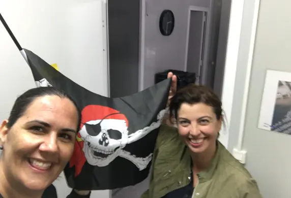 Two smiling ladies indoors holding up a flag with a skull in a red bandana and cross bones on it