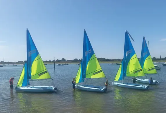 A group of Fusion sail boats being held by the sailors at the shore edge with bright blue and yellow mainsails and jibs up on a sunny day