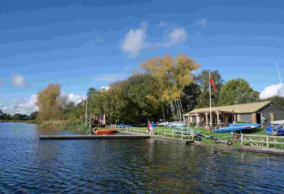 A club house with jetty into the water at the front with trees in the background on a clear sunny day