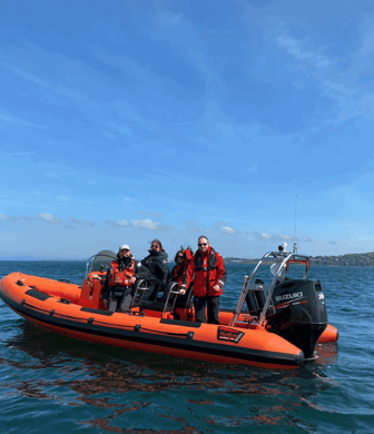 Five people stood on an orange rigid inflatable boat (RIB) on clear blue water on a sunny day