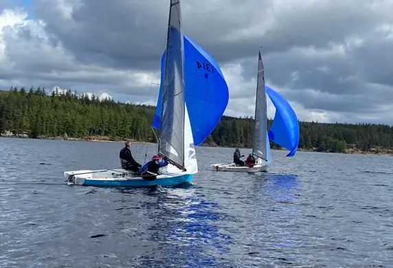 Two people sailing dinghies with main sails, jibs and blue spinnakers up