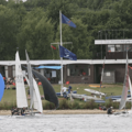 A variety of sailing dinghies on the shoreline with the club house and trees in the background