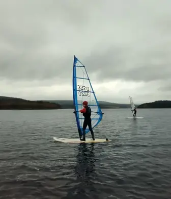 A person stood on a windsurf with another in the distance on flat calm water on a grey cloudy day