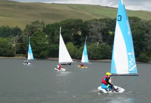 Four pico sailing dinghies on the water with trees and hills in the background on a cloudy day