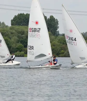 Laser dinghies racing in a line one behind the other on calm water with trees on the shore in the background