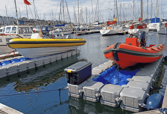 One yellow and one orange rigid inflatable boat (RIB) sitting on a floating dock in a mariner with lots of masts in the background