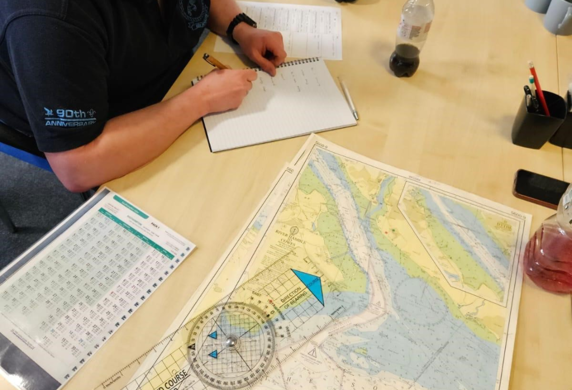A student sat at a desk surrounded by navigational charts and equipment making notes in a notebook at Ocean Sports Tuition, Southampton